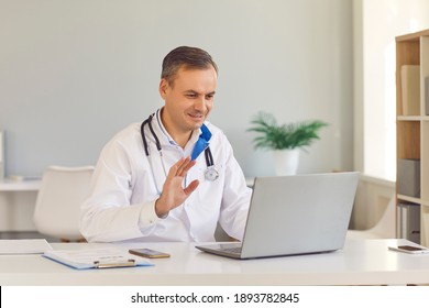 Smiling Man, Online EHealth Service Doctor, Sitting At Desk In Office And Waving Hand At Laptop Computer, Saying Hello To Patient During Video Call. Telemedicine And Virtual Visit To Hospital Concept