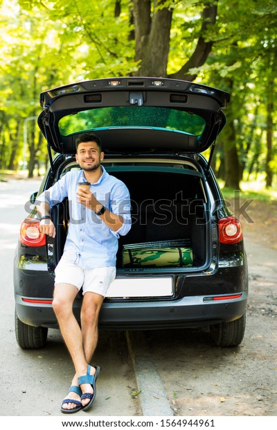 smiling man with paper cup\
of coffee sitting in car trunk and looking at camera while using\
smartphone