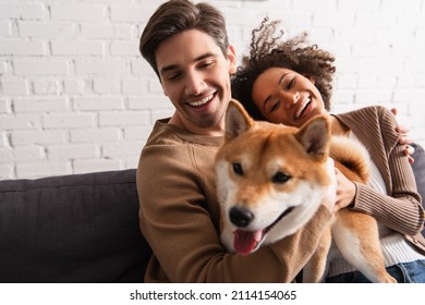 Smiling man petting blurred shiba inu dog near african american girlfriend on couch - Powered by Shutterstock