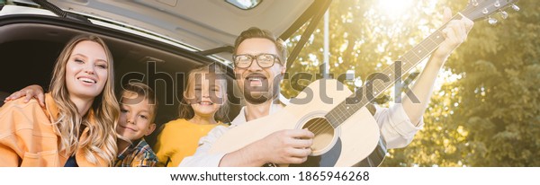 Smiling man playing acoustic guitar near family in car\
trunk, banner 