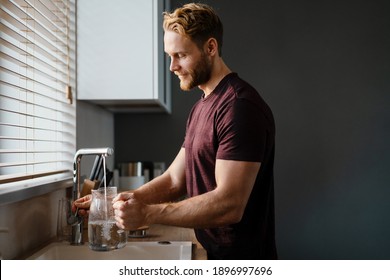 Smiling man pouring water in the jug from a tap while standing at the kitchen sink - Powered by Shutterstock