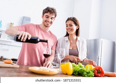 Smiling man pouring wine near girlfriend with knife and vegetables on blurred foreground - Powered by Shutterstock