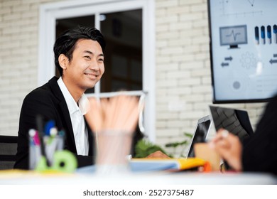 A smiling man in a professional setting, engaged in conversation with a colleague, surrounded by office elements and a screen displaying data visuals. - Powered by Shutterstock