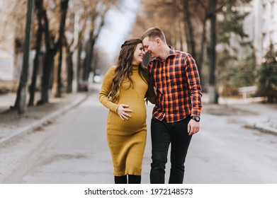 A smiling man in a red plaid shirt and a pregnant woman in a yellow dress are walking along the street in nature. Pregnancy photography.