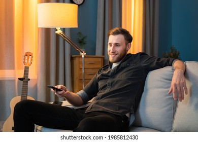 A Smiling Man Is Relaxing After Work In His Homey, Cozy Living Room In The Evening. Boy Is Sitting On The Sofa, Leaning Against The Cushion, And Holding The TV Remote In His Hand, Changing Channels