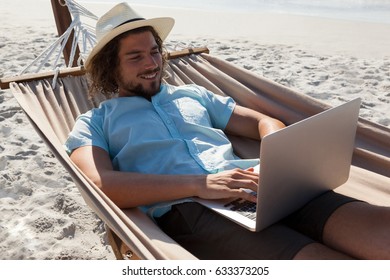 Smiling Man Relaxing On Hammock And Using Laptop On The Beach