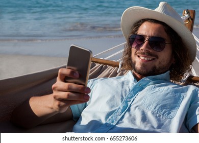 Smiling Man Relaxing On Hammock And Using Mobile Phone On The Beach