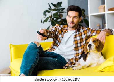 Smiling Man With Remote Control Watching Tv And Sitting On Sofa With Dog 