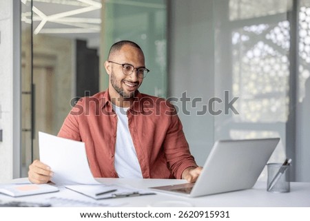 A smiling man reviews documents while working on his laptop in a modern office setting, suggesting remote work or online collaboration.