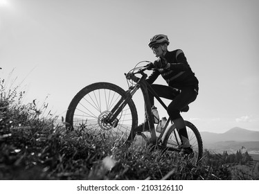 Smiling man in safety helmet and glasses cycling uphill on sunny day. Male bicyclist in cycling suit climbing uphill on mountain bike. Concept of sport and active leisure. Monochrome image. - Powered by Shutterstock