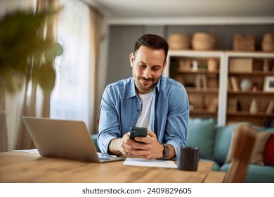 A smiling man scrolling through social media and checking his messages on his cellphone while sitting at a table with a laptop in front of him. - Powered by Shutterstock