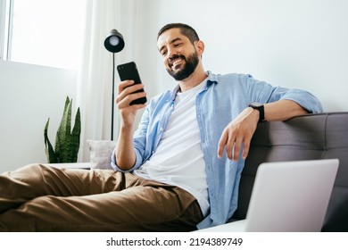 Smiling man sitting on couch at home using laptop and cell phone - Shutterstock ID 2194389749