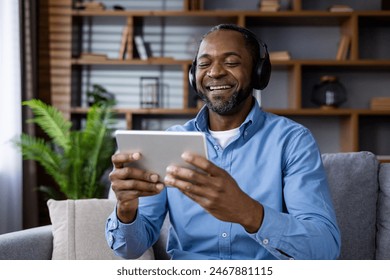 Smiling man sitting on a couch wearing headphones, watching something on his digital tablet in a living room setting. Shelves with decor items and a plant in the background. - Powered by Shutterstock