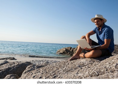 Smiling Man Sitting On The Rocks And Using Laptop On Beach