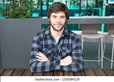 Smiling man sitting at street cafe table with his arms crossed. Young guy wearing casual shirt and sitting at wooden table with cafe interior in background. Young man portrait concept. Front view.