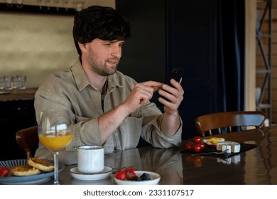 Smiling man sitting at a table in the kitchen with a cup of coffee and looking at a smartphone. A man with glasses enjoys breakfast.  - Shutterstock ID 2331107517
