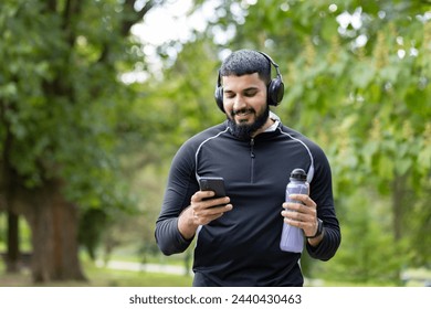 Smiling man in sportswear holding a water bottle while using his smartphone and wearing headphones in a park setting. - Powered by Shutterstock