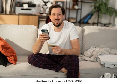 A Smiling Man With Stubble Is Sitting On A Couch In Pajamas, Next To A Pizza Box, A Mess, A Man Is Holding A Phone And A Credit Card, He Is Shopping Online, Ordering Food Through An App.