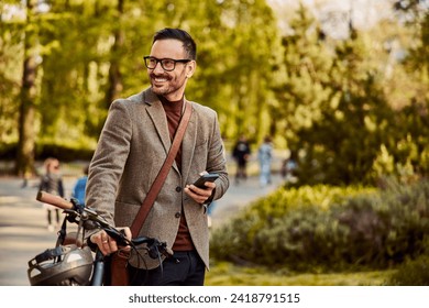 A smiling man in a suit pushing his bicycle, holding a mobile phone, and looking around the park during the walk home from work. - Powered by Shutterstock