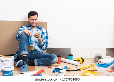 Smiling Man Surfing Internet On Mobile Phone While Sitting Near Wall. House Remodeling And Interior Renovation. Young Bearded Guy Sitting On Floor Among Cardboard Boxes And Painting Tools At Home.