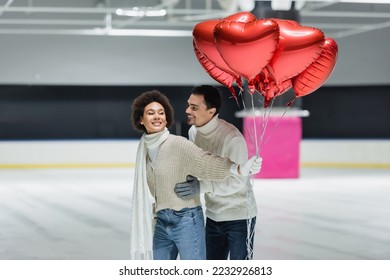 Smiling man in sweater hugging african american girlfriend with balloons in heart shape on ice rink  - Shutterstock ID 2232926813