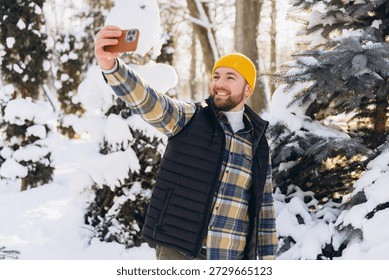 Smiling man taking a selfie while enjoying winter weather in a snowy park, wearing a yellow beanie, plaid shirt, and vest