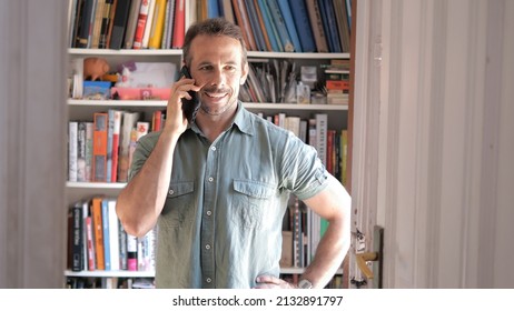 Smiling Man Talking On Cell Phone In A Room With Bookcase Behind. Selective Focus