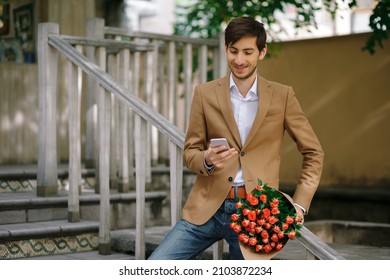 Smiling man texting while looking at phone and holding bunch of roses, folded in craft paper, and standing near wooden railing outdoors, wearing jeans, wool jacket, white shirt. Confident man.