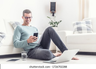 Smiling Man Texts On His Mobile Phone During His Home Office Hours At Home, Sitting On A Living Room Floor.