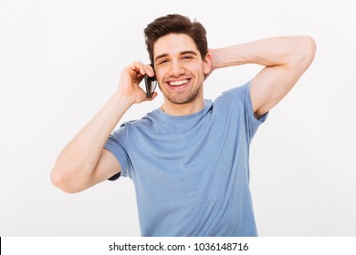 Smiling Man In T-shirt Talking By Smartphone While Holding His Head And Looking At The Camera Over Grey Background