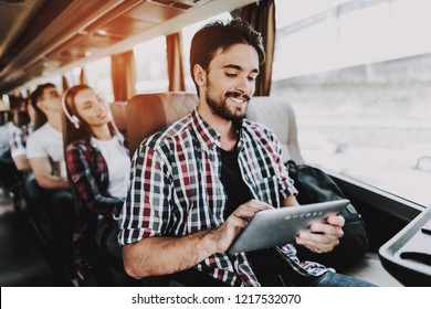 Smiling Man Using Digital Tablet In Tourist Bus. Young Handsome Man Sitting On Passenger Seat Of Tour Bus And Holding Touchscreen Device. Traveling And Tourism Concept. Happy Travelers On Trip