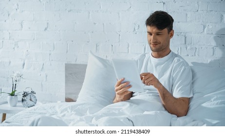 smiling man using digital tablet while sitting in bed near alarm clock on bedside table