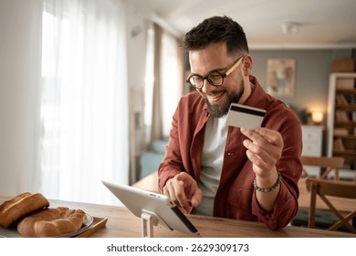 Smiling man using a digital tablet and credit card for online payment, enjoying a secure and convenient shopping experience in his modern apartment