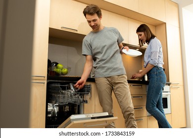 Smiling Man Is Using Dishwasher While Young Woman Cooking Healthy Breakfast On The Background