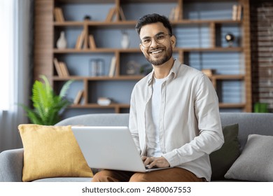 Smiling man using laptop while sitting on couch. Modern living room with bookshelf in background. Concept of remote work, technology, and happiness.
