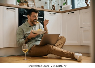 Smiling man using laptop while eating spaghetti at home. Copy space.