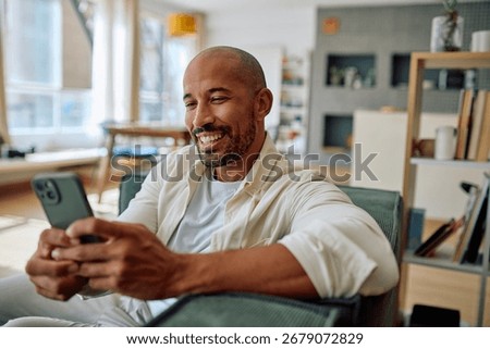 Smiling man using mobile phone while sitting on comfortable sofa in living room, enjoying online communication or social media