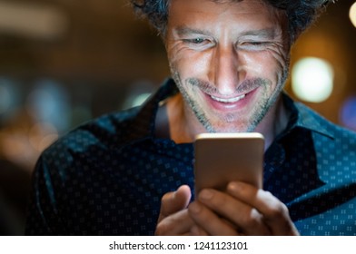 Smiling Man Using Smart Phone During Night. Closeup Face Of Happy Mature Businessman Messaging On Cellphone At Night On The Street With The Lights Of The Road Blurred In The Background.