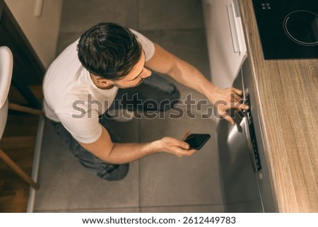 Smiling man using smartphone to control and turn on dishwasher in his kitchen at home.