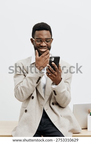 Smiling man using smartphone, dressed in beige blazer and glasses, in bright modern office with laptop on desk, casual business attire, cheerful expression, technology lifestyle concept, indoor