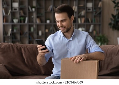 Smiling Man Using Smartphone, Holding Cardboard Box, Unpacking Parcel Browsing Mobile Device Apps, Blogger Shooting Unboxing Video, Happy Customer Satisfied By Good Delivery Service, Sitting On Couch