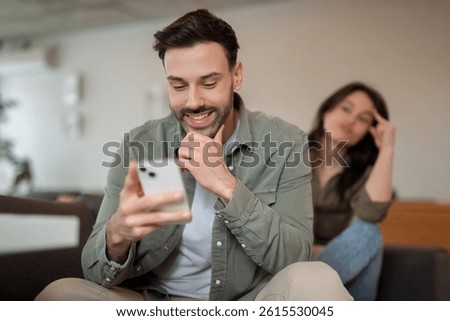 Smiling man using smartphone while a frustrated woman sits in the background, looking annoyed and disappointed.