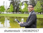 Smiling man using tablet and standing in city park. Guy wearing casual clothes and holding gadget with pond and green trees in background. Communication and nature concept. Side view.