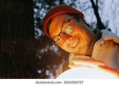 Smiling man wearing glasses and a red wool hat holds a glowing sphere in a forest at night, creating a mysterious and magical atmosphere - Powered by Shutterstock