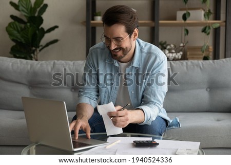 Smiling man wearing glasses using laptop, calculating domestic bills, planning, managing budget, sitting on couch at home, happy businessman browsing online banking service, satisfied by money refund