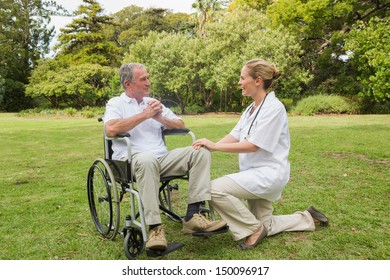 Smiling Man In A Wheelchair Talking With His Nurse Kneeling Beside Him In The Park On Sunny Day