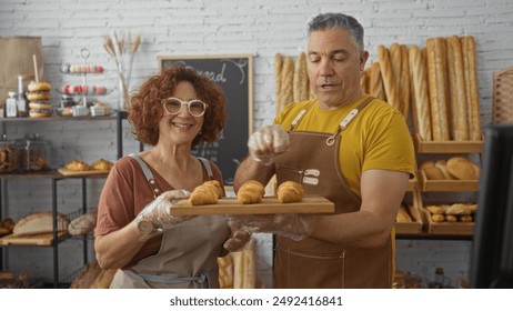 Smiling man and woman bakers working together in a bakery holding a tray of fresh croissants in an indoor shop setting with breads and pastries displayed in the background - Powered by Shutterstock