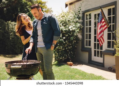 Smiling Man And Woman Cooking In Their Backyard With American Flag In The Background. Couple Making  Grilled Food Celebrating An Occasion.