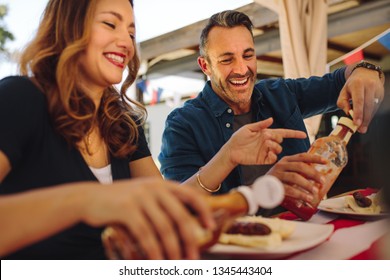 Smiling Man And Woman Eating Snacks Sitting At A Restaurant. Happy Couple Pouring Tomato Sauce On Hot Dogs Sitting At A Dining Table.