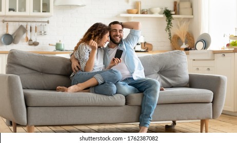 Smiling man and woman hugging using smartphone reading joke and laughing sitting on couch in living room. Loving husband embracing young wife holding smartphone in hand watching funny video at home. - Shutterstock ID 1762387082